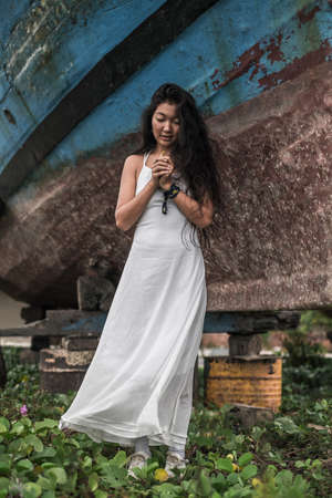 Charming young mongol woman in white dress looking at her hands clenched at the chest. Brunette long...