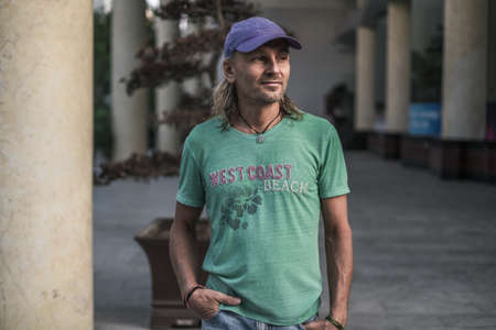 Mature man with long gray hair wearing green t-shirt and blue cap standing on the street in town and...