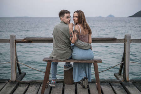 Beautiful young couple in love sitting on the bench by the sea, cuddling and looking at camera with copy space. High quality photoの写真素材
