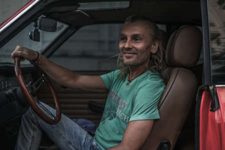 Portrait of mature handsome man sitting in red old style car, holding steering wheel and smiling....