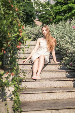 Pretty girl in casual white dress sitting on the stairs in the park. Long blond hair. Romantic summer or spring pictureの写真素材