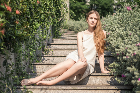 Beautiful young caucasian woman in white dress sitting on the stairs in the park. Long blond hair. High quality photoの写真素材