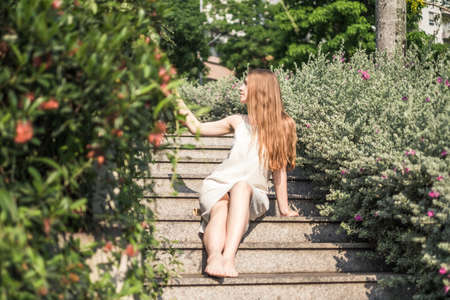 Beautiful young caucasian woman in white dress sitting on the stairs in the park and looking at flowers. Long blond hair. Beautiful plants.の写真素材