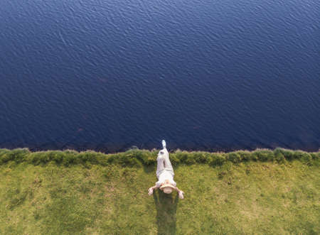 Picture created by dji camera. Young woman in white dress sitting on the grass on the shore edge near the sea. Up side view. Blue seaの写真素材