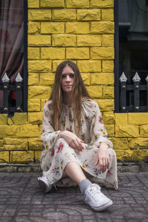 Pretty hipster caucasian young woman in casual dress and sneakers sitting on the street. Long straight blond hair. Pensive lookの写真素材