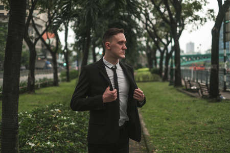 Portrait of handsome caucasian young businessman in black elegant suit walking in the park. Looking aside. Serious look. Summer time.の写真素材