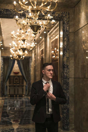 Portrait of young handsome business man in elegant business suit walking on the hall with beautiful expensive chandeliers. straightens his tieの写真素材