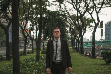 Portrait of young man in elegant business suit walking in summer street, outdoors. Looking at camera with copy space. Green parkの写真素材