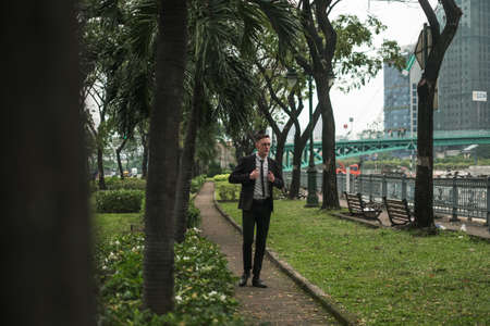 Attractive young caucasian man in black formal suit walking in the park. Park with tropical plants. High quality photoの写真素材