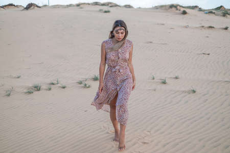 Beautiful blond woman in long dress and jewellery on her head walking in desert with white sand on sunny day during summer vacation. Sand dunes.の写真素材