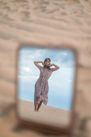 Photo in the mirror of attractive blond woman in long dress walking in desert. Wearing jewellery on her head. Picture in the mirror. Sand dunes.の写真素材