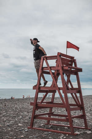 Man stands on a rescue tower with the red flag on the dark pebble beach in Batumi, Georgia. Looking at the sea. High quality photoの写真素材
