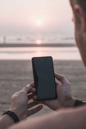 Over shoulder view: man hands holding black smartphone with blank screen on the beach. Mock up, template, entertainment and technology conceptの写真素材