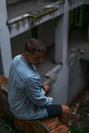 Portrait of young handsome Caucasian man in denim jacket, sitting on the abandoned construction site and looking downの写真素材