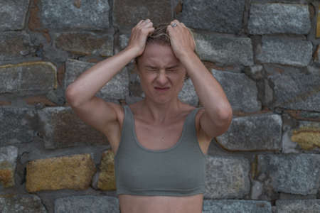 Portrait of stressful angry young caucasian blonde woman in crop top on the stone background. Angry girl tears the hair on her head. panic attacksの写真素材