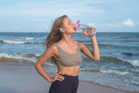 Close up Portrait of sporty fit pretty young blonde caucasian woman drinking water on the beach after sport training. reusable sports bottle, oceanの写真素材