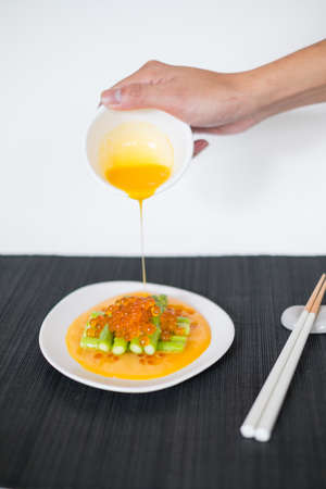 Hand pours sauce on mini asparagus with red caviar. White plate and chopsticks on black mat in Japanese restaurant, close up, picture for the menuの写真素材