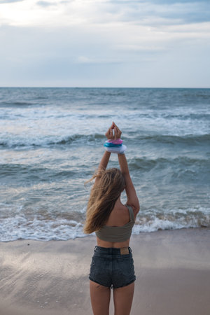 Blond young woman with hands wrapped in blue, pink and white plastic bags on the beach. Back view....