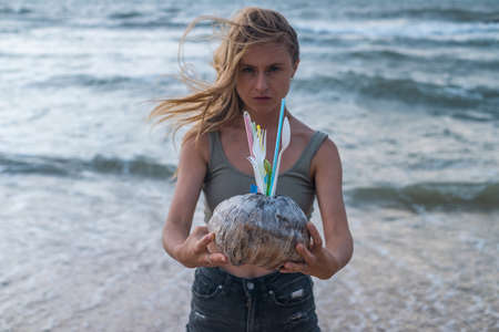 Sad blond caucasian young woman standing on the beach and holding old coconut with used plastic dishes and straws. Sea on the background.の写真素材
