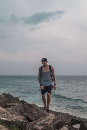 Young man in cap, t-shirt and shorts walks on stones by the sea. Full arm tattoo. Windy day. Traveller walks near the ocean.の写真素材