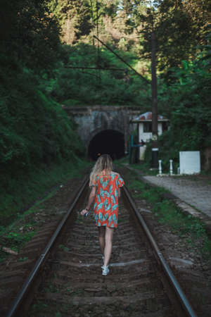 Young woman in colourful dress walks along the rails in the forest. Lonely girl approaches a tunnel...