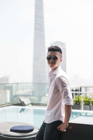Handsome young Asian man standing in the rooftop bar near the swimming pool. Chinese guy, millennials life, lifestyle in the big city, Modern Asiaの写真素材
