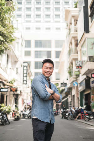 Portrait of asian young handsome man in denim shirt standing on the street with crossed hands, smiling and looking at camera with copy space.の写真素材
