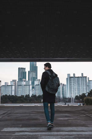 Young man in a hat, black coat and jeans with backpack walks on the street in metropolis. skyscrapers on the background. traveller in big city.の写真素材