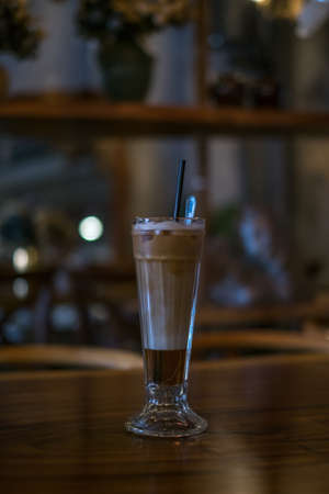 Close-up photo of glass cup with ice latte on the wooden table in a coffee shop. Glass with iced caramel cappuccino with the straw on the tableの写真素材