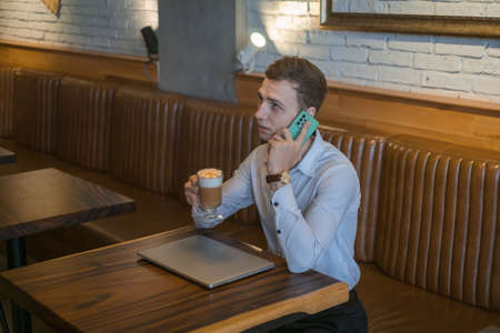 Young business man talking on mobile phone while drinking coffee in a bar. Freelancer working at a cafe. Caucasian man speaks on the phoneの写真素材