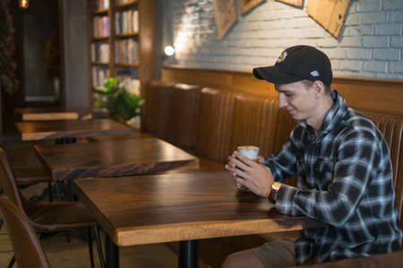 Smiling young caucasian man in checkered shirt and cap drinks coffee in a bar. Coffee shop, cafe,...