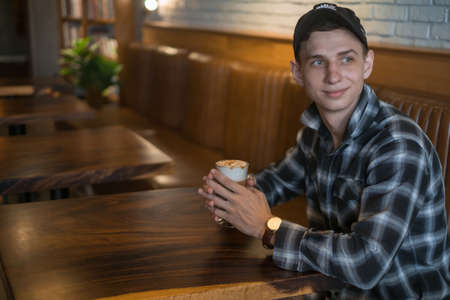 Smiling young caucasian man in checkered shirt and cap drinks coffee in a bar. Coffee shop, cafe, latte, caramel cappuccino. High quality photoの写真素材