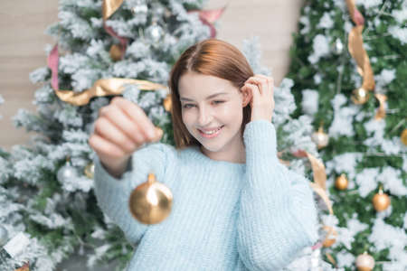 New Year portrait of beautiful caucasian young woman in cozy wool warm light blue sweater holding a gold ball for decoration christmas tree.の写真素材