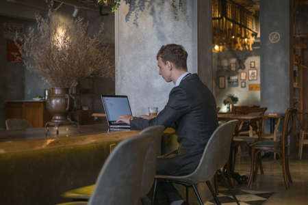 Young caucasian business man in formal suit working on laptop while drinking coffee in a bar. Handsome man working at a cafe, typing on keyboardの写真素材