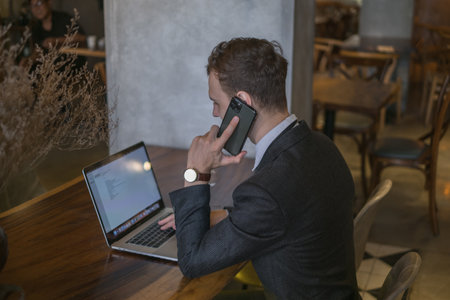 Young business man talking on mobile phone while drinking coffee and working with laptop in a bar. Caucasian handsome man working at a cafeの写真素材