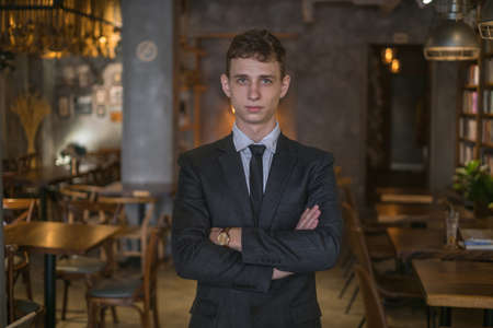 Portrait of young caucasian business man in formal suit standing in the bar with crossed arms. Handsome man standing in the coffee shopの写真素材