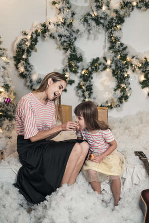 Christmas studio family shoot. New Years photo of Beautiful Mother and cute daughter have fun together. Mom feeds her daughter with a tangerineの写真素材