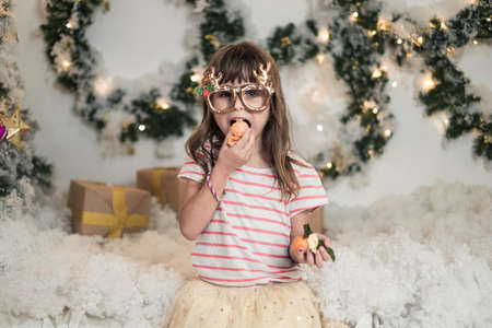 Christmas studio shoot. New Years photo of happy cute girl wearing funny Christmas glasses and eating mandarin. Holding a tangerine in his handの写真素材