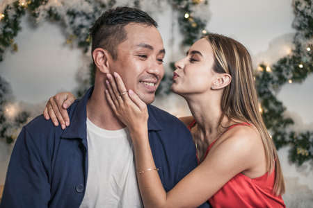Close up portrait of lovely couple. New Years photo of happy couple. Christmas wreaths on the background. Woman kissing man on his cheekの写真素材