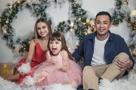 Christmas studio family shoot. New Years photo of happy family. Christmas wreaths and artificial snow. Mother, father and cute surprised daughter.の写真素材