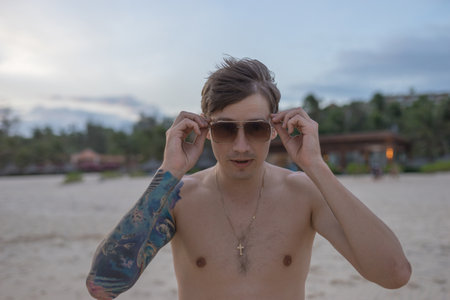 Portrait of handsome caucasian young man standing on the beach and wearing sunglasses, full arm tattoo. high quality photoの写真素材