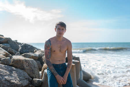 Handsome young caucasian bare-chested man with full-arm tattoo sitting on the rock by the sea. high quality photoの写真素材