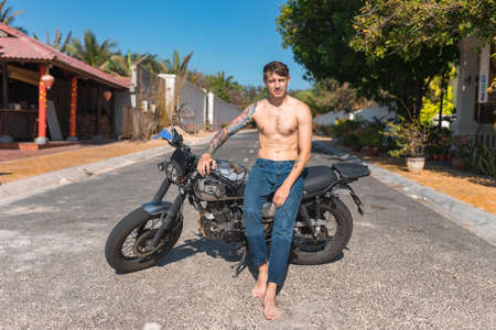 Young shirtless caucasian man with full-arm tattoo sitting on motorcycle on the street. Handsome fit bare-chested guy sits on moto. high quality photoの写真素材