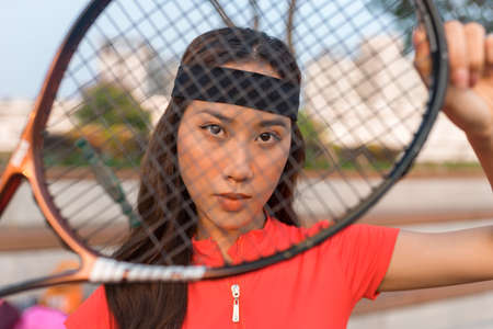 Asian young woman with black headband standing on the tennis court and holding tennis racket. High...