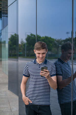 Young caucasian man in casual clothing standing near modern glass building and looking at his phone. People using phones. High quality photoの写真素材