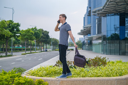 Young caucasian man in casual clothing with the backpack walking near modern office buildings and making phone call. people using phone. High qualityの写真素材