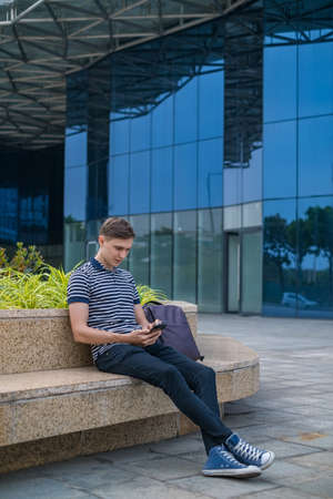 Young caucasian man in casual clothing sitting near modern office building and looking at his phone. High quality photoの写真素材