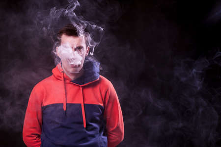A young dark-haired man with sad eyes in a colored orange and blue hoody standing with arms behind...
