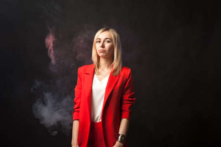 Young beautiful white blond girl in a bright red strict suit with a jacket and white blouse looks at the camera on a background of colored blue and pink smoke on a black isolated backgroundの写真素材