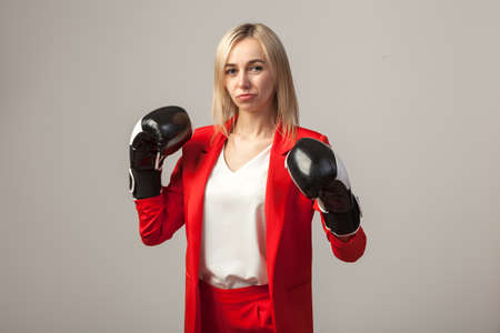 Young beautiful white blond girl in a bright red strict suit with a jacket and white blouse standing in a pose with boxing gloves on a white isolated backgroundの写真素材